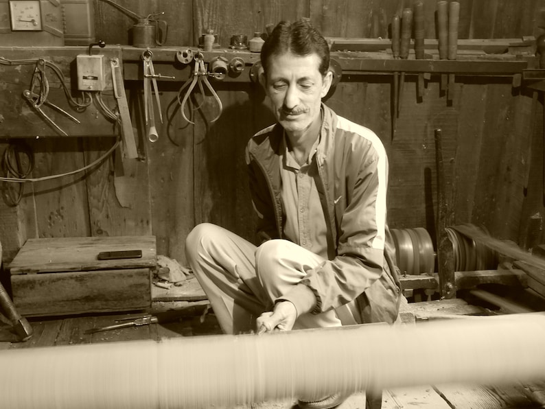 May include: A sepia-toned image of a man working in a workshop. He is seated, focused on a spool of thread or yarn. The workshop has wooden walls and various tools hanging on the wall.