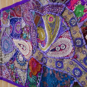 Op de afbeelding: Een kleurrijk patchwork textiel met diverse patronen en texturen. Het is overwegend paars, blauw en goud en bevat paisley- en bloemmotieven, verfraaid met pailletten en gedetailleerde stiksels. De randen zijn afgewerkt met een paarse rand.