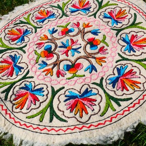 Op de afbeelding: Een ronde, crèmekleurige vloerkleed met een levendig bloemenpatroon. Het vloerkleed heeft een centraal bloemmotief, omringd door een ring van bloemen en bladeren. De bloemen zijn in blauw, oranje, rood en groen, met een rode zigzagrand.