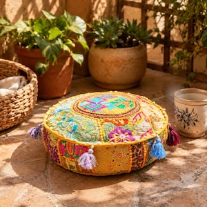 May include: A round, yellow floor cushion featuring a patchwork design with intricate multi-colored embroidery, floral patterns, and geometric shapes. The cushion is accented with small fabric tassels in shades of blue, purple, and pink.