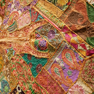 May include: Close-up of a vibrant, patchwork textile. The design features a rich tapestry of colors including gold, red, purple, and green. Intricate patterns, including paisley and floral motifs, are visible throughout the fabric. The textile appears to be hand-stitched.