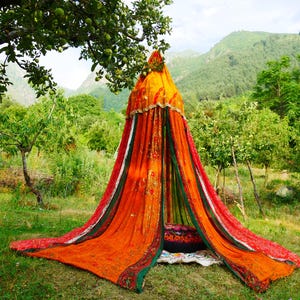 May include: A conical fabric canopy featuring vibrant orange, red, and green panels with intricate gold-toned embroidery and decorative trim. The structure drapes down to the ground, creating a tent-like enclosure with a dark circular cushion inside.