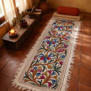 Puede incluir: Una alfombra rectangular de color blanco roto con un intrincado patrón de bordado floral en tonos rosa, azul, amarillo y verde. La alfombra presenta un fleco grueso y deshilachado a lo largo de los bordes largos y un borde cosido.