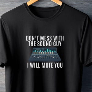 Pode incluir: Camiseta preta com o texto "DON'T MESS WITH THE SOUND GUY I WILL MUTE YOU". O design inclui um gráfico de uma mesa de mistura em azul, verde e branco.