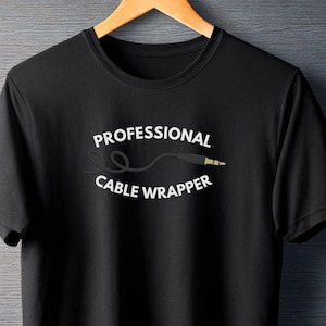 以下が含まれることがあります： 黒いTシャツ。白文字で「PROFESSIONAL CABLE WRAPPER」とケーブルとジャックのグラフィックがプリントされています。木製のハンガーに掛けられ、暗い背景に配置されています。