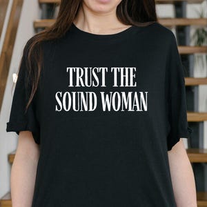 Pode incluir: T-shirt preto com o texto branco "TRUST THE SOUND WOMAN". A camisa é de gola redonda e parece ser feita de um material macio. A pessoa está usando jeans cinza.