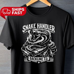 Pode incluir: Camiseta preta com um gráfico branco de uma cobra enrolada em cabos de áudio. O texto "SNAKE HANDLER" está acima da cobra, com "BACKLINE CO." e "LIVE SOUND STAGE CREW" abaixo.