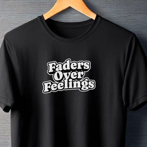 Pode incluir: T-shirt preto com o texto branco "Faders Over Feelings" em uma fonte retrô. A camiseta é exibida em um cabide de madeira contra um fundo cinza. A camiseta de manga curta é feita de um material macio.