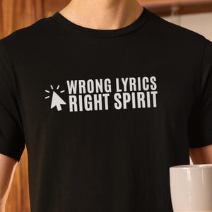 Pode incluir: Camiseta preta com o texto branco "WRONG LYRICS RIGHT SPIRIT" e um cursor. A pessoa está segurando uma caneca branca e bege. A camisa é de gola redonda.