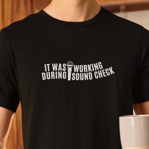 Pode incluir: Camiseta preta com o texto branco "IT WAS WORKING DURING SOUND CHECK" e um gráfico de microfone. A camiseta é de gola redonda. Uma pessoa está segurando uma caneca branca e bege.