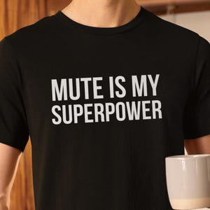 Pode incluir: Camiseta preta com gola redonda com o texto branco "MUTE IS MY SUPERPOWER". A pessoa está segurando uma caneca branca e bege. A camisa é feita de um material macio.