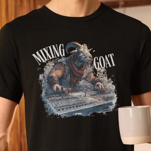 Pode incluir: Camiseta preta com um desenho de uma cabra usando uma mesa de mistura. O texto "MIXING GOAT" está acima da ilustração da cabra. A pessoa na imagem está segurando uma caneca branca.