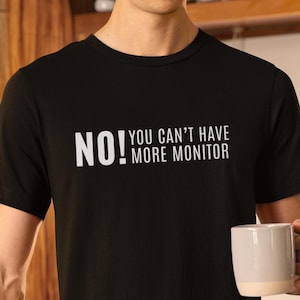 May include: Black crew neck t-shirt with the text "NO! YOU CAN'T HAVE MORE MONITOR" in white. The person is holding a white and beige mug.