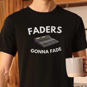 Pode incluir: T-shirt preto com o texto branco "FADERS GONNA FADE". A camisa apresenta uma ilustração de uma mesa de mistura de som. A pessoa na imagem está segurando uma caneca branca.