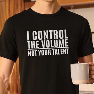 Pode incluir: Camiseta preta com o texto branco "I CONTROL THE VOLUME NOT YOUR TALENT". A camisa é de manga curta. Uma pessoa está segurando uma caneca branca.
