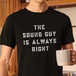 Pode incluir: Camiseta preta com o texto branco "THE SOUND GUY IS ALWAYS RIGHT". A camiseta é de gola redonda. Uma pessoa segura uma caneca branca.