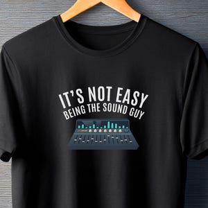 Pode incluir: Camiseta preta com o texto branco "IT'S NOT EASY BEING THE SOUND GUY". Abaixo, um gráfico de uma mesa de som com luzes azuis e verdes. A camiseta está pendurada em um cabide de madeira.