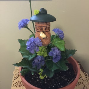 May include: A small terracotta pot with a green birdhouse on top, surrounded by blue flowers and green leaves. The birdhouse is made from a cork with the words "You're My Little" written on it. A small white bird is perched inside the birdhouse.