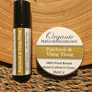 Op de afbeelding: Een klein, wit potje met een gouden deksel en etiket. Het etiket luidt "Organic Natural Deodorant Patchouli & Ylang Ylang 100% Plant Based Hand Crafted in Essex 35ml e".  Naast het potje staat een klein, bruin glazen rolletje met een zwarte dop en een etiket met de tekst "Aromatherapy Remedy Oil Patchouli & Ylang Ylang 100% Natural - Alcohol Free Emotionally Uplifting & Balancing Anxiety. Aphrodisiac"
