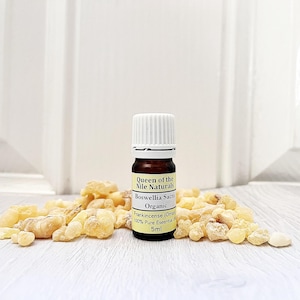 Könnte beinhalten: Eine kleine braune Glasflasche mit weißem Verschluss, beschriftet mit "Queen of the Nile Naturals" und "Frankincense (Oman) 100% Pure Essential Oil 5ml." Die Flasche ist von hellfarbigen, unregelmäßig geformten Harzstücken umgeben.