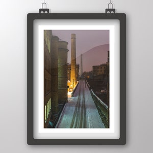 Può includere: Una fotografia di un edificio in mattoni con un alto camino in fondo. L'edificio è ricoperto di neve e la strada di fronte è vuota. Il cielo è di un colore viola chiaro.