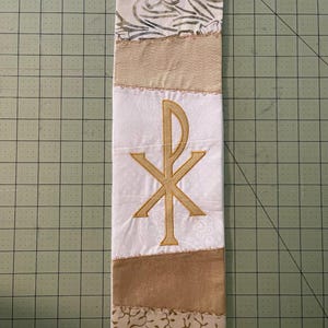 May include: A white and gold liturgical stole with a Chi Rho symbol embroidered in gold thread. The stole features various fabric panels with floral and patterned designs, and gold trim.