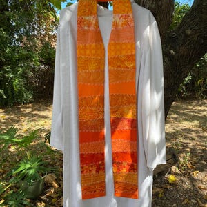 Könnte beinhalten: Eine lange, orangefarbene und gelbe Stola aus Stoff mit Patchwork-Muster. Die Stola hängt an einem Kleiderbügel vor einem weißen Hintergrund.
