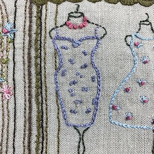 Puede incluir: Obra de arte bordada que representa una escena de boutique con vestidos en exhibici&oacute;n. Los vestidos est&aacute;n delineados en azul y adornados con peque&ntilde;os detalles florales. Un collar rosa adorna el maniqu&iacute;. El fondo tiene l&iacute;neas verticales en varios tonos de marr&oacute;n y verde.