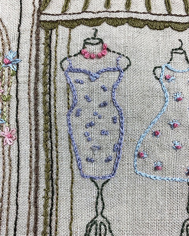 Puede incluir: Obra de arte bordada que representa una escena de boutique con vestidos en exhibici&oacute;n. Los vestidos est&aacute;n delineados en azul y adornados con peque&ntilde;os detalles florales. Un collar rosa adorna el maniqu&iacute;. El fondo tiene l&iacute;neas verticales en varios tonos de marr&oacute;n y verde.