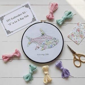 Puede incluir: Kit de bordado DIY con diseño de pez de rayos X. Incluye un bastidor, hilos de colores, tijeras y un paquete de agujas con estampado floral. El texto en la tarjeta dice "DIY Embroidery Kit - 'X' is for X-Ray Fish."