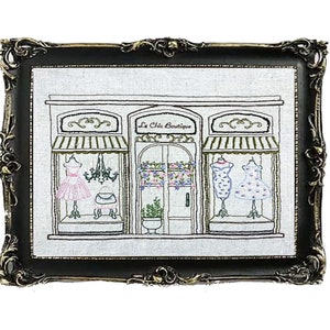 Puede incluir: Obra de arte bordada de un escaparate de boutique, enmarcada en un marco negro y dorado ornamentado. La boutique presenta un letrero que dice "Le Chic Boutique" y exhibe vestidos en los escaparates. La obra de arte est&aacute; sobre un fondo blanco.