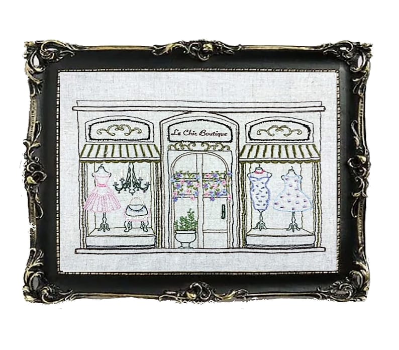 Puede incluir: Obra de arte bordada de un escaparate de boutique, enmarcada en un marco negro y dorado ornamentado. La boutique presenta un letrero que dice "Le Chic Boutique" y exhibe vestidos en los escaparates. La obra de arte est&aacute; sobre un fondo blanco.