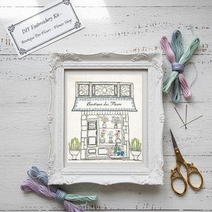 May include: A framed DIY embroidery kit featuring a floral shop design. The white frame surrounds the artwork, with spools of thread in pastel colors, gold scissors, and needles. The kit includes the text "DIY Embroidery Kit - Boutique Des Fleurs - Flower Shop."