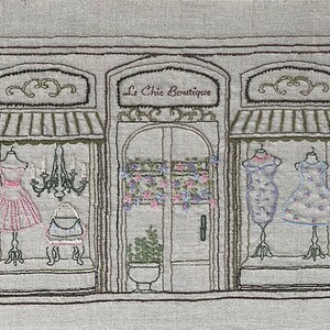 Puede incluir: Imagen bordada de un escaparate de una boutique, con el texto "Le Chic Boutique". La tienda exhibe vestidos en maniqu&iacute;es en los escaparates. Una l&aacute;mpara de ara&ntilde;a cuelga en el interior y detalles florales adornan la puerta y las ventanas, creando una escena encantadora y detallada.