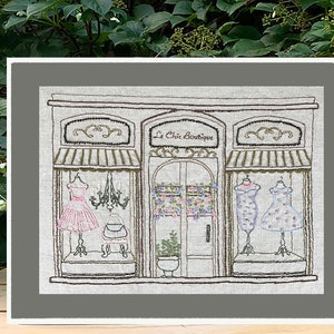 Puede incluir: Una obra de arte bordada representa un escaparate de una boutique con el texto "Le Chic Boutique". La tienda presenta tres escaparates con vestidos en maniqu&iacute;es, una l&aacute;mpara de ara&ntilde;a y detalles florales. La obra est&aacute; enmarcada en un color neutro.