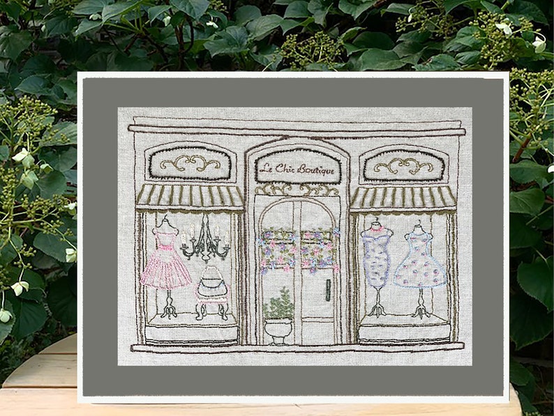 Puede incluir: Una obra de arte bordada representa un escaparate de una boutique con el texto "Le Chic Boutique". La tienda presenta tres escaparates con vestidos en maniqu&iacute;es, una l&aacute;mpara de ara&ntilde;a y detalles florales. La obra est&aacute; enmarcada en un color neutro.