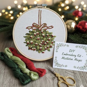 Könnte beinhalten: Ein DIY-Stickset mit einem Mistelzweig-Design. Das Set enthält einen Stickrahmen mit einem gestickten grünen und roten Mistelzweig, Stickgarn in Grün und Rot, eine goldene Schere und eine Karte mit "DIY Embroidery Kit - Mistletoe Magic".