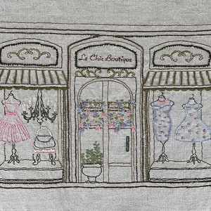 Puede incluir: Imagen bordada de un escaparate de boutique, "Le Chic Boutique", con vestidos en maniqu&iacute;es en los escaparates. La tienda tiene un toldo y adornos marrones, con una l&aacute;mpara de ara&ntilde;a y detalles florales. Los vestidos son rosas, azules y blancos.