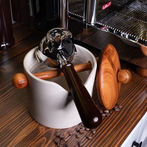 Könnte beinhalten: Ein verchromter Espressomaschinen-Siebträger ruht in einem cremefarbenen Keramikhalter mit Holzgriff. Ein Holz-Tamper und Kaffeebohnen sind ebenfalls auf einer dunklen Holzoberfläche zu sehen. Die Espressomaschine befindet sich im Hintergrund.