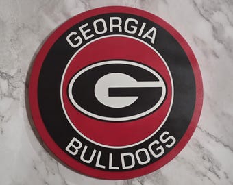 May include: A round sign featuring the Georgia Bulldogs logo. The sign has a red outer ring, a black inner ring, and a white and black central design with the letter "G". The words "GEORGIA" and "BULLDOGS" are in white text.