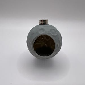 May include: A gray, spherical flask with a textured surface resembling the moon. The flask has a large opening and a silver cap. The interior appears gold. A unique and decorative item.