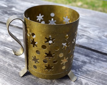 Puede incluir: Un portavasos de metal dorado con un diseño decorativo de estrellas y círculos recortados. El portavasos tiene un asa y tres patas.