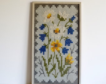 Puede incluir: Un bordado de punto de cruz enmarcado que presenta un diseño floral con flores blancas, amarillas y azules sobre un fondo blanco.