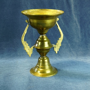 Puede incluir: Un cáliz de latón antiguo con un cuenco ancho y poco profundo y una base acampanada. El cáliz presenta asas decorativas con un diseño calado estilizado. El color general es un dorado cálido y envejecido, lo que sugiere que es una pieza antigua o vintage.