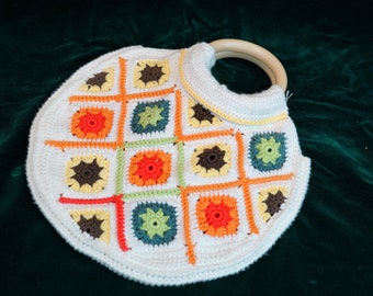 May include: A white crocheted bag with a wooden handle. The bag is decorated with colorful squares in shades of orange, yellow, green, and brown.