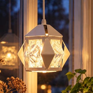 Op de afbeelding: Een witte geometrische hanglamp met een gefacetteerde, heldere glazen kap in een wit metalen frame. De lamp verspreidt een warme gloed en verlicht de complexe, hoekige structuur van de lamp die aan een wit snoer hangt.