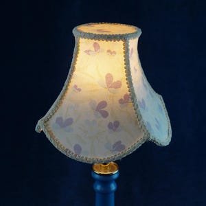 K&ouml;nnte beinhalten: Ein wei&szlig;er Lampenschirm mit einem Blumenmuster und goldenen Verzierungen. Der Lampenschirm ist beleuchtet und das Licht scheint durch den Stoff.