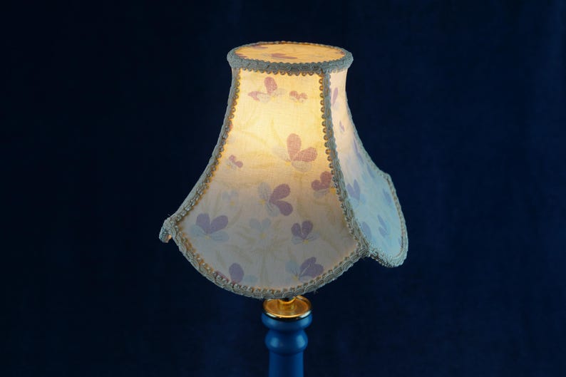 K&ouml;nnte beinhalten: Ein wei&szlig;er Lampenschirm mit einem Blumenmuster und goldenen Verzierungen. Der Lampenschirm ist beleuchtet und das Licht scheint durch den Stoff.
