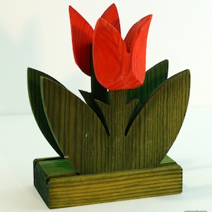 May include: Wooden napkin holder shaped like a tulip with red flowers and green leaves. The holder has a base for holding napkins.