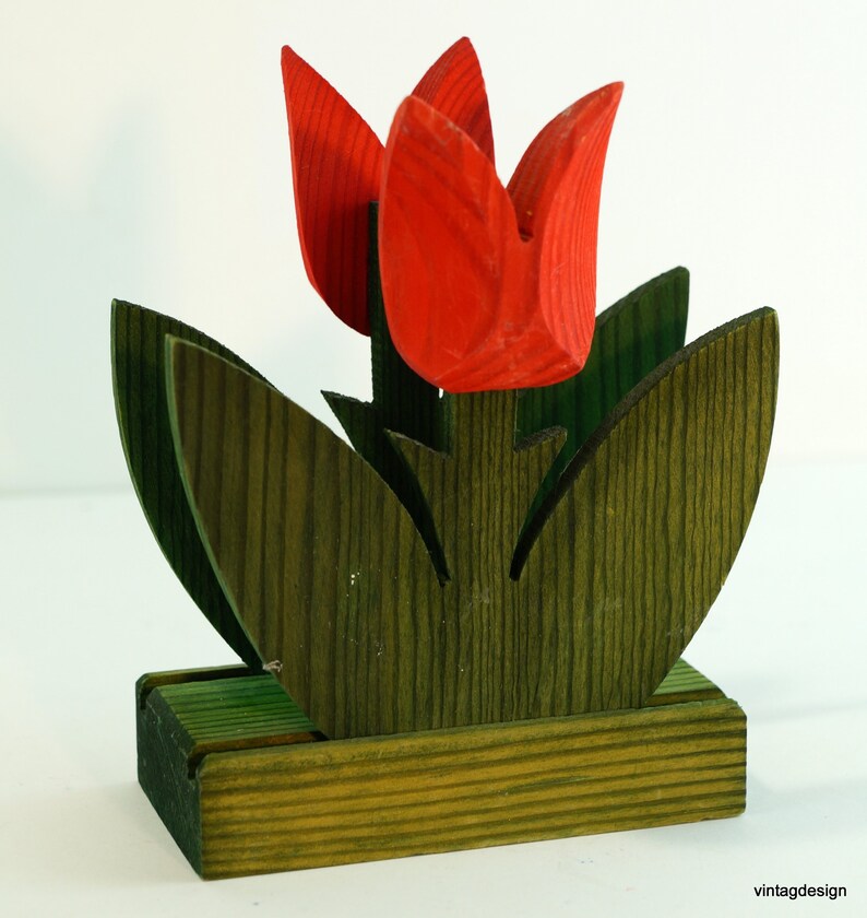 May include: Wooden napkin holder shaped like a tulip with red flowers and green leaves. The holder has a base for holding napkins.